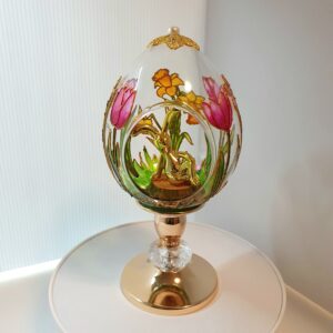 Handmade glass Easter egg on stand with stained glass painting of tulips and daffodils, yellow bunny sitting on a stump inside, decorative floral filigree on top
